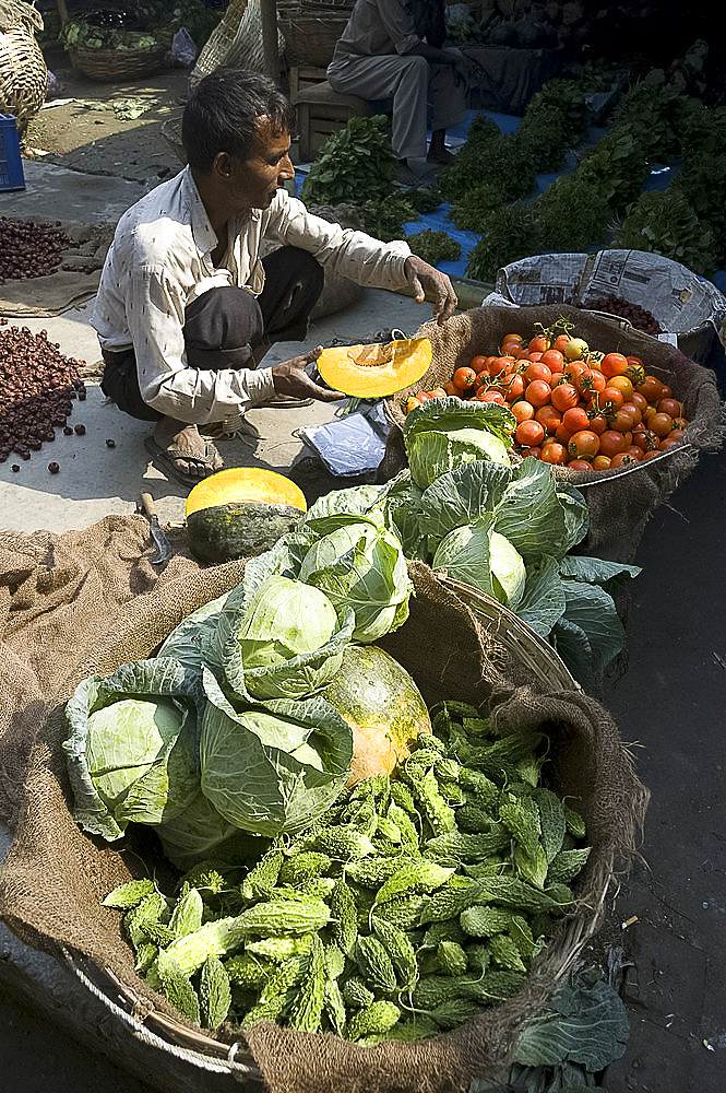 Vegetable wallah preparing pumpkin with baskets of tomatoes, cabbages and bitter gourd in early morning market on the banks of the Brahmaputra river, Guwahati, Assam, India, Asia