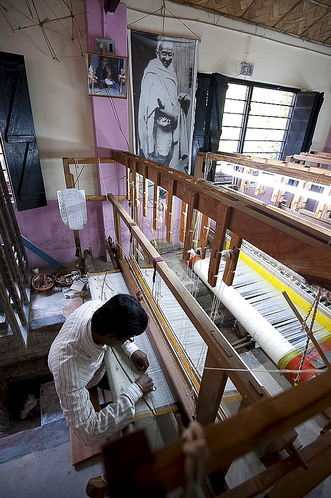 Man working a loom in a village weaving workshop beneath a large picture of Mahatma Gandhi, Kalna, West Bengal, India, Asia