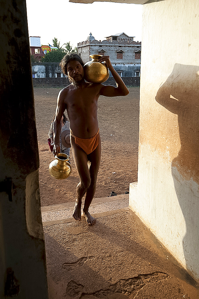 Monk, follower of Mahima Dharma sect, wearing orange loincloth carrying brass pots of water into the temple at sunset, Joranda, Orissa, India, Asia