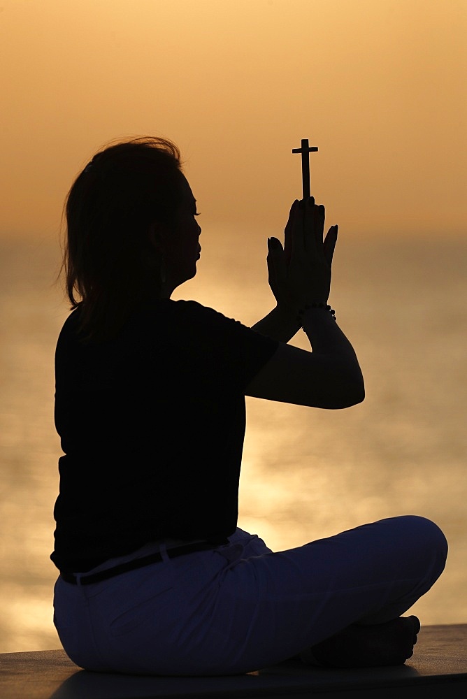 Silhouette of faithful woman praying with Christian cross at sunset as concept for religion, faith, prayer and spirituality, Dubai, United Arab Emirates, Middle East