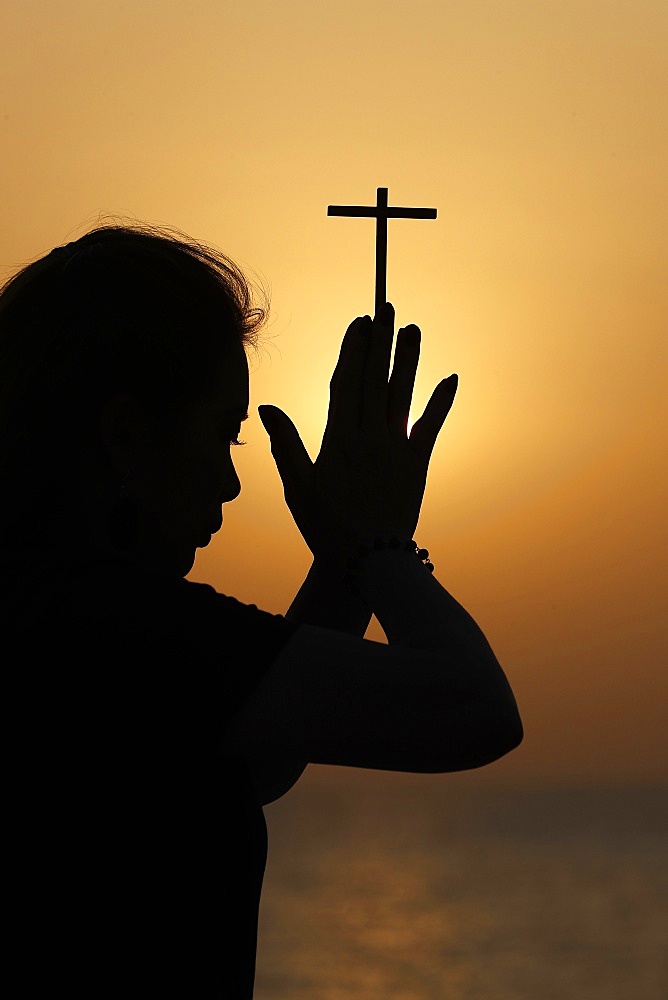 Silhouette of faithful religious woman praying with Christian cross at sunset, United Arab Emirates, Middle East