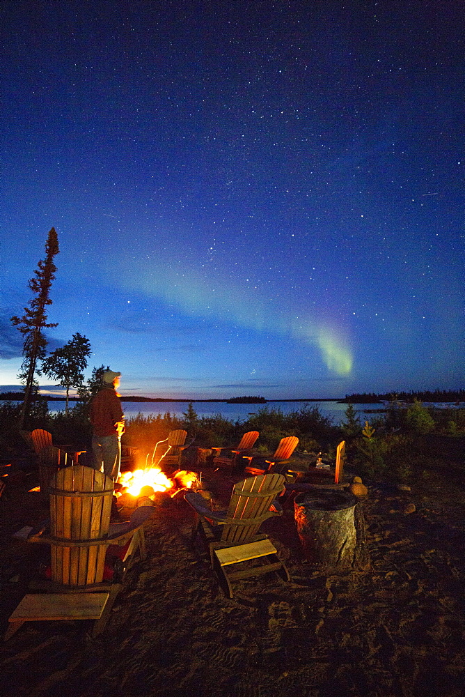 A man views the Northern Lights (aurora borealis) by a camp fire by Lake Egenolf in northern Manitoba, Canada, North America