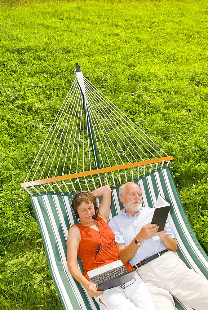 Senior citizen couple, woman wearing a headset looking at a netbook or laptop, man reading a book