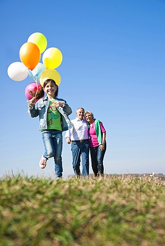 Girls holding balloons in her hand while running across a meadow in front of her grandparents