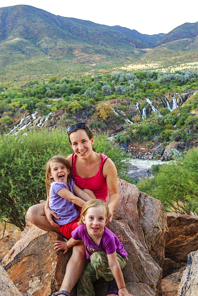 Mother with two girls sitting on rocks in green landscape, behind Epupa Falls, Kunene, Kunene region, Namibia, Africa