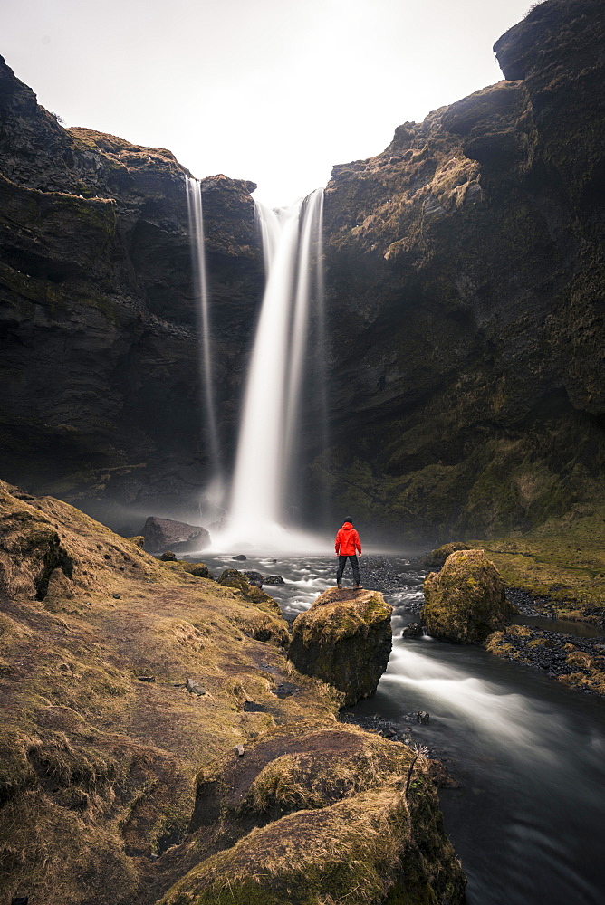 Man in red jacket in front of Kvernufoss waterfall in a gorge, dramatic atmosphere, time exposure, near Skógafoss, Southurland, Iceland, Europe