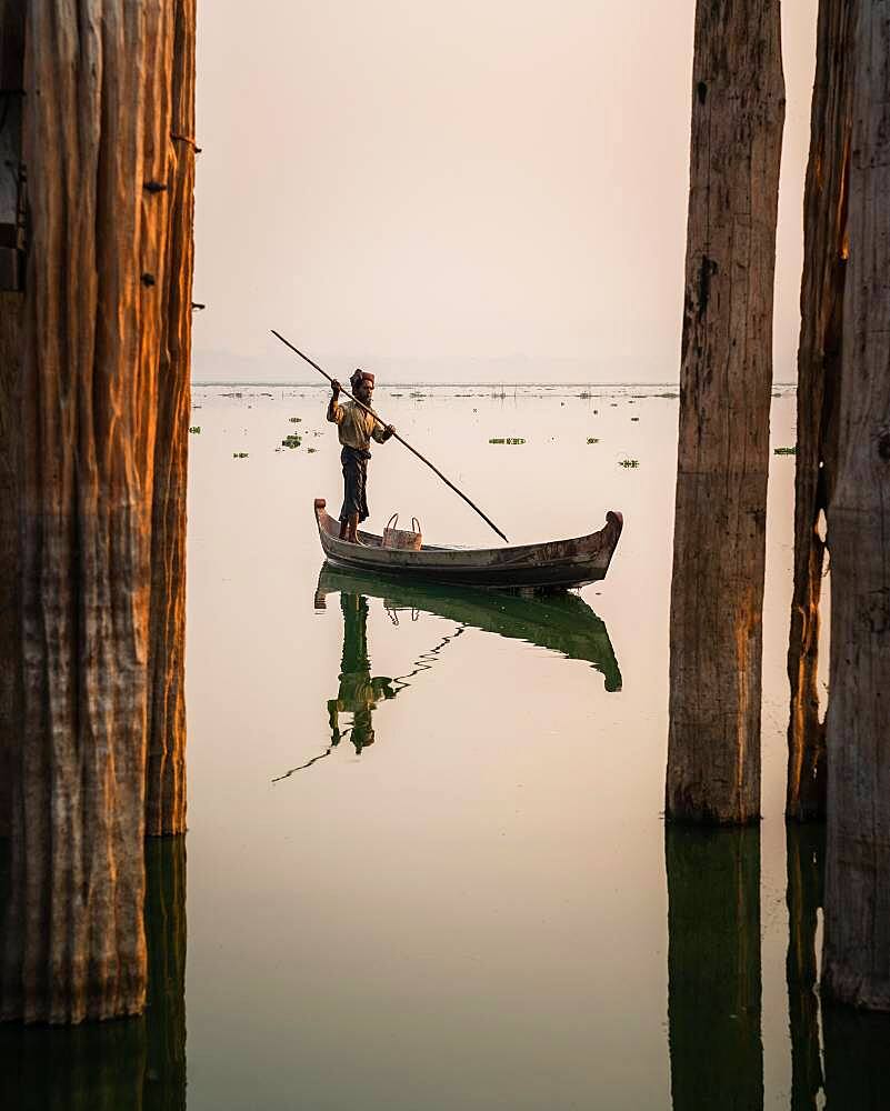 Fisherman stands with boat on Taung Tha Man Lake for sunrise between the posts of the U-leg bridge, reflection in the water, Thaung Tha Man Lake, Mandalay, Myanmar, Asia