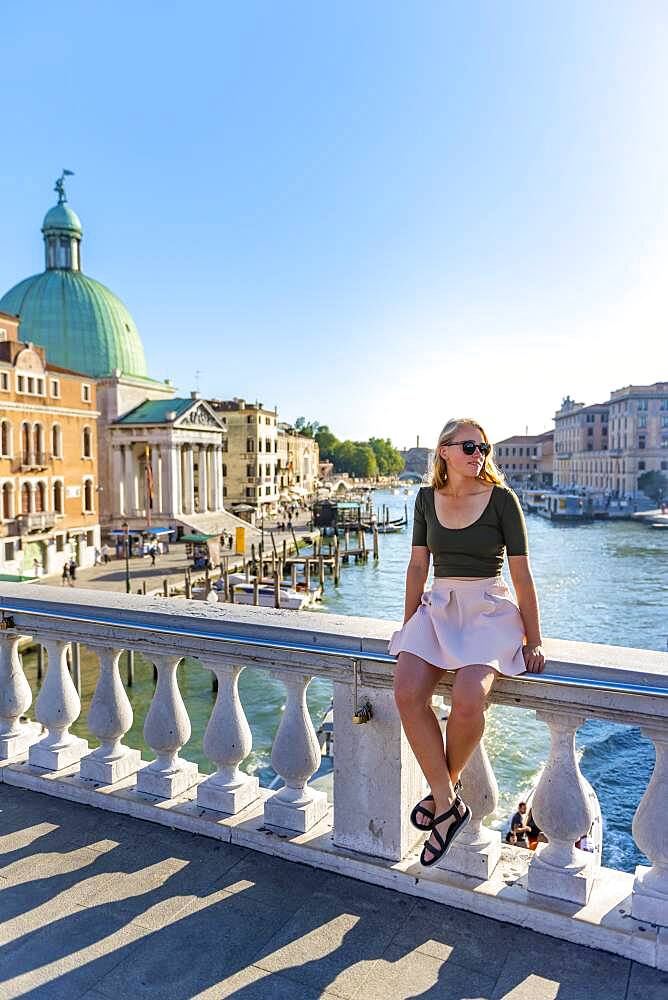 Young woman sitting on bridge railing on the Grand Canal, Church of San Simeone Piccolo, Ponte degli Scalzi, Venice, Veneto, Italy, Europe