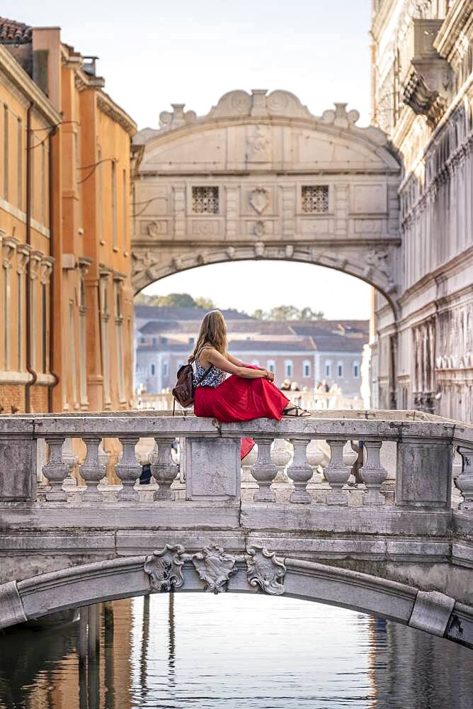 Young woman with red skirt, tourist sitting on a bridge railing, bridge over the Rio di Palazzo, behind Bridge of Sighs, Venice, Veneto, Italy, Europe