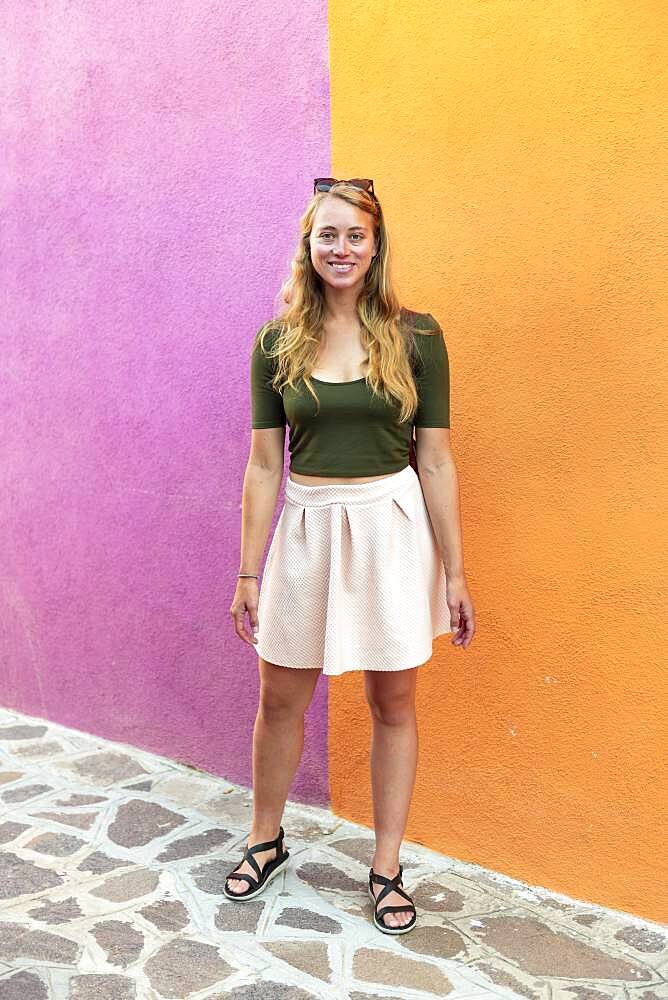 Young woman in dress stands laughing in front of colorful house, orange and pink house facade, Burano Island, Venice, Veneto, Italy, Europe