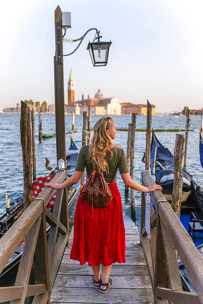 Young woman with red dress on a jetty, Venetian gondolas, in the back church San Giorgio Maggiore, Venice, Veneto, Italy, Europe