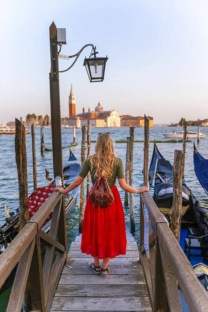 Young woman with red dress on a jetty, Venetian gondolas, in the back church San Giorgio Maggiore, Venice, Veneto, Italy, Europe