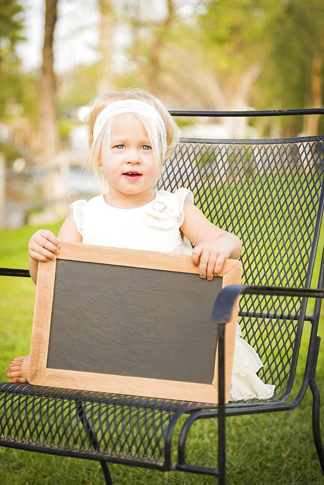 Cute baby girl sitting in chair holding blank blackboard