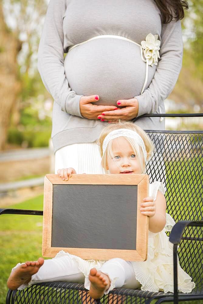 Pregnant mom behind cute baby girl sitting in chair holding blank blackboard