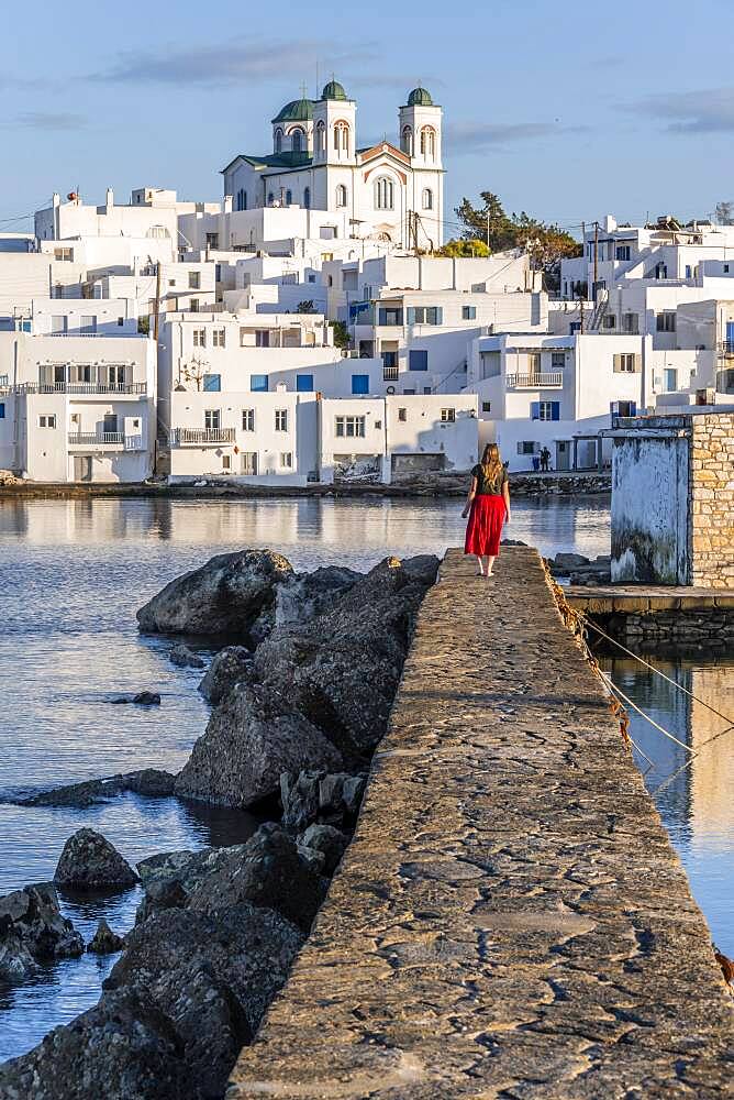 Young woman with red dress on harbour wall, behind church of Naoussa, harbour town Naoussa, island Paros, Cyclades, Greece, Europe