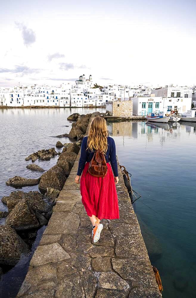 Young woman with red dress on harbour wall, behind church of Naoussa, harbour town Naoussa, island Paros, Cyclades, Greece, Europe