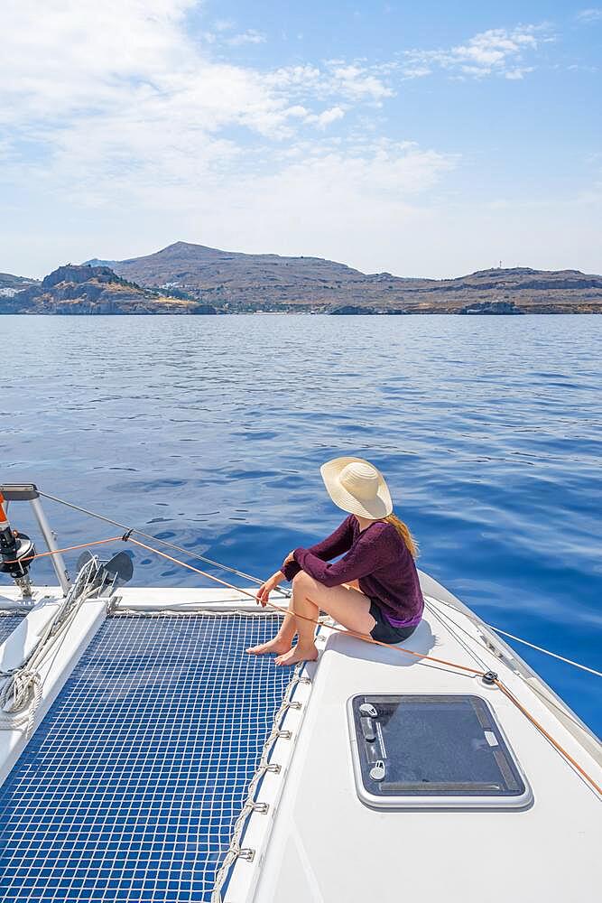 Young woman with hat sitting on deck at the net of a sailing catamaran, sailing trip, Rhodes, Dodecanese, Greece, Europe