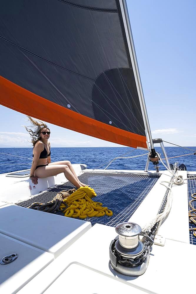 Young woman in bikini sitting in the net of a sailing catamaran, headsail and ropes sailing, sailing trip, Dodecanese, Greece, Europe