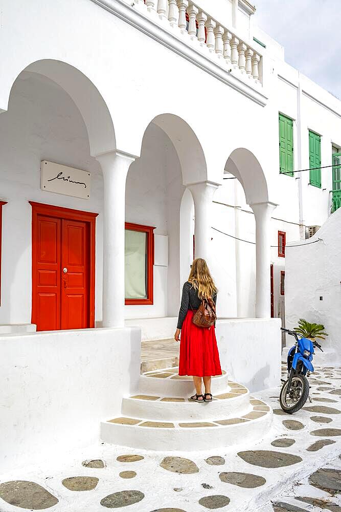 Young woman in red skirt in front of a white Cycladic house with columns and red door, alleys of the old town Chora, Mykonos Town, Mykonos, Cyclades, Greece, Europe