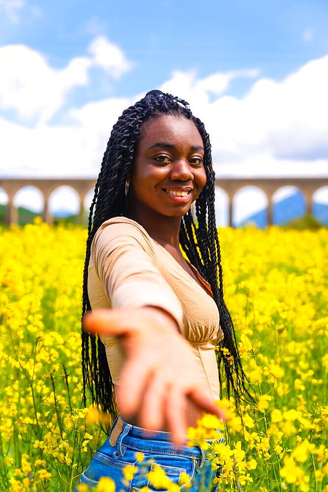 Lifestyle, offering the hand, portrait of a black ethnic girl with braids, traveler, in a field of yellow flowers