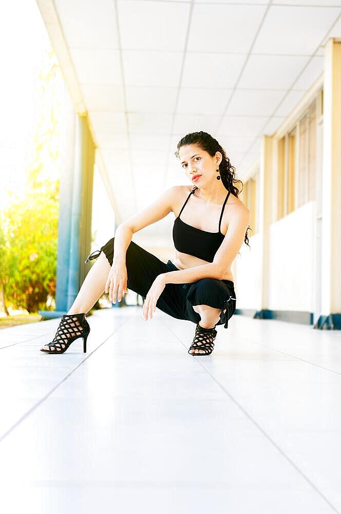 Dance artist woman in high heels outdoors. Portrait of dance girl in high heels crouching on the floor. Portrait of dancer Woman in heels on the floor looking at the camera