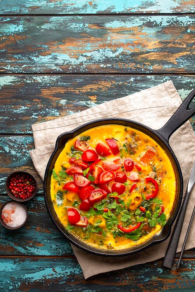 Healthy frittata in cast iron pan with fried beaten eggs and seasonal vegetables on rustic wooden background. Italian omelette with organic spinach, bell pepper, tomatoes, top view