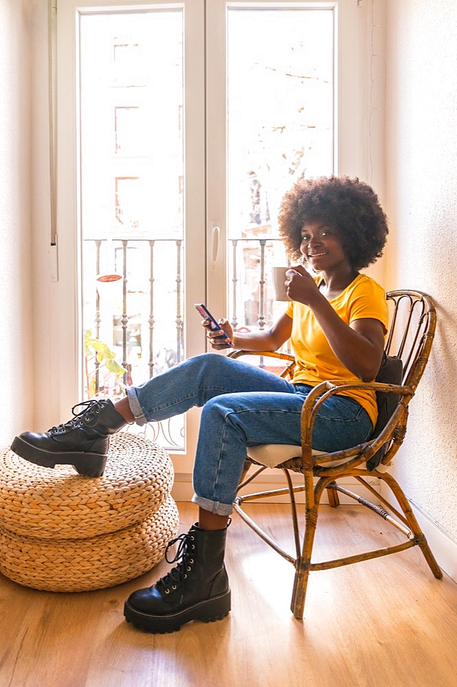 Afro hair woman by the window home having a hot drink, coffee in the morning looking at the mobile