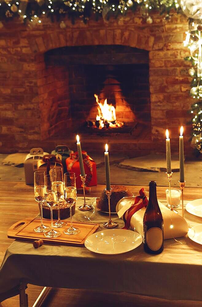 Christmas or New year dinner against burning fireplace and xmas gifts in cozy house decorated with led lights, festive table with freshly baked cake, lit candles, glasses of champagne, selective focus