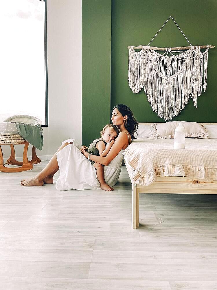 Young woman mother expressing love to little baby, sitting on the floor by bed with sleeping child in hands and kissing him in forehead, tender photo of unconditional love between mom and son