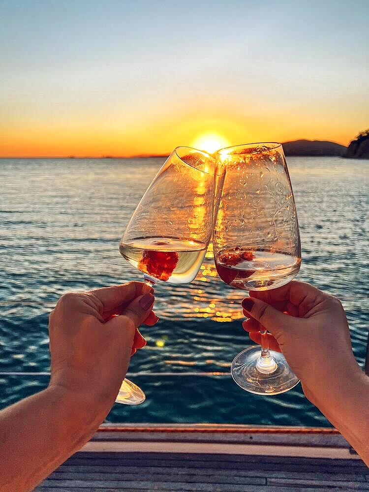 Female hands holding glasses of champagne with raspberries on board the boat. Sunset sky and sea on the background. Making a celebratory toast with sparkling wine. Birthday, holiday, party and friendship concept