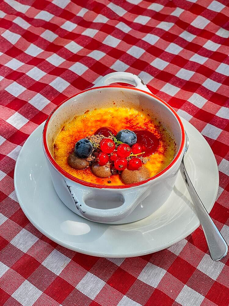 Creme brulee, burnt cream. Sweet baked dessert in a deep porcelain white plate with handles. Dish, decorated with currants and blueberries. Plate and spoon are placed on a red checkered tablecloth