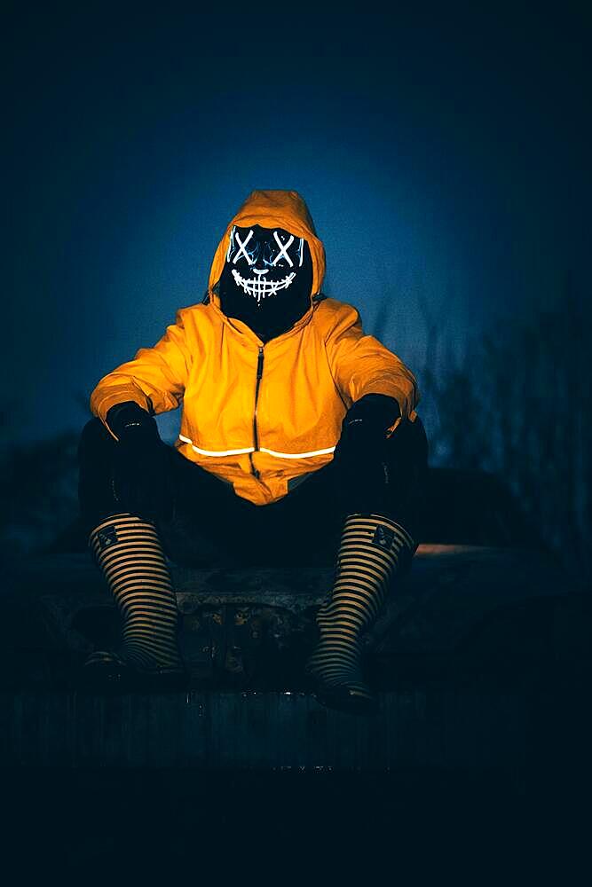 Purge mask, man with LED mask on a burnt out car, rubber boots and yellow rain jacket, Frankfurt am Main, Hesse, Germany, Europe