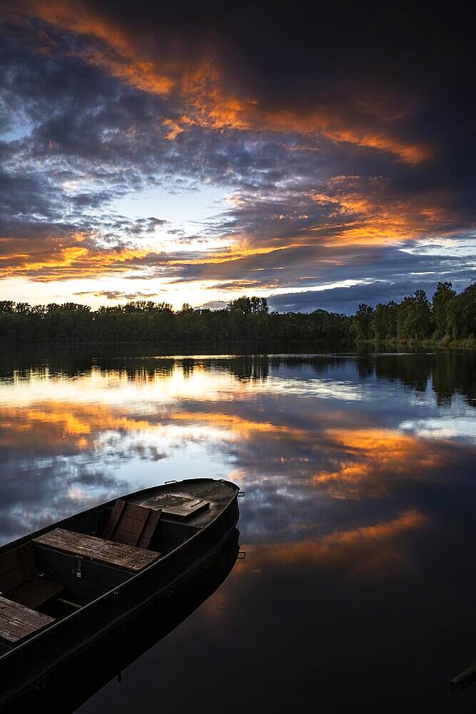 A small boat in a lake at sunset with coloured clouds in orange in the sky. The sky is reflected in the calm lake. Rhine-Neckar District, Baden-Wuerttemberg, Germany, Europe