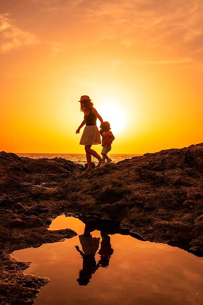 Silhouette of mother and son walking in the sunset on the beach of Tacoron in El Hierro, Canary Islands, orange sunset, walking along the sea pointing the path