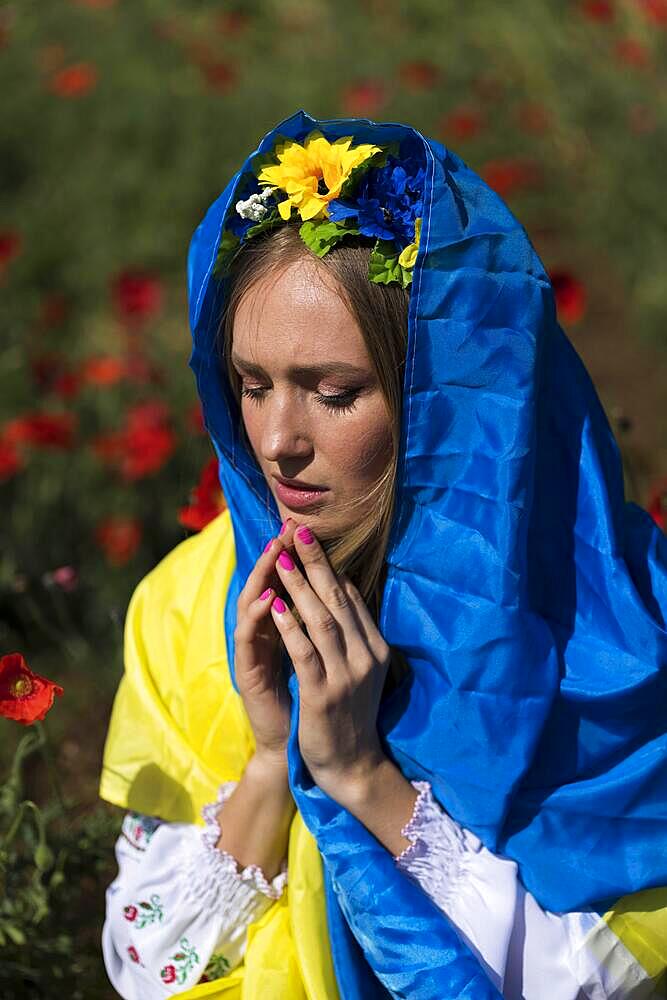 A young blonde Ukrainian woman stands in a field of Red Poppy flowers holding the flag of Ukraine showing her support for the war in her native country of Ukraine