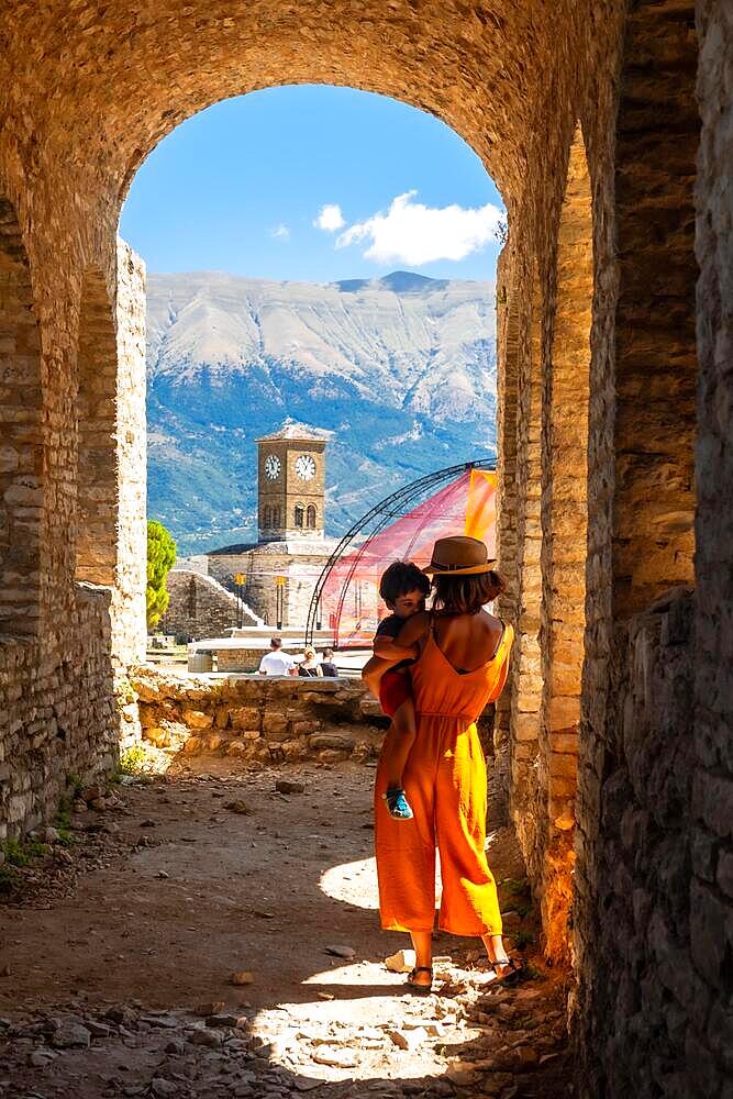 A woman with her son walking in the arches of the fortress of the Ottoman castle of Gjirokaster or Gjirokastra and in the background the church with the clock tower. Albania, Kulla e Sahatit