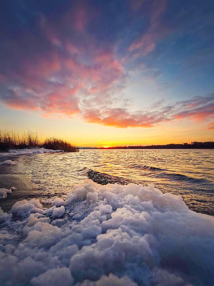 Windy evening at the lake with waves creating white foam on the shore. Vibrant sunset scene over the pond