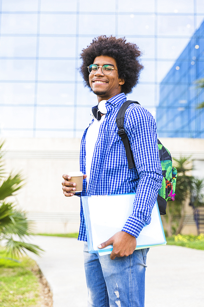 Portrait young male student holding disposable coffee cup books hand standing against campus