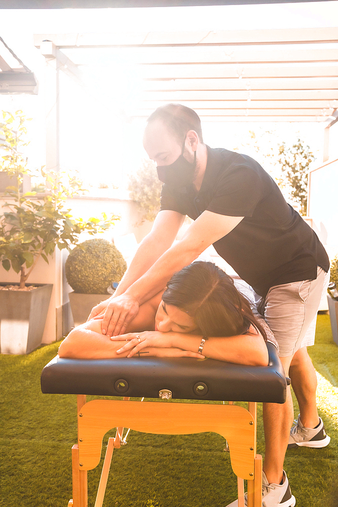 Massage at the client's home on the terrace, photo at sunset. Masseur with face mask in the coronavirus pandemic, vertical photo