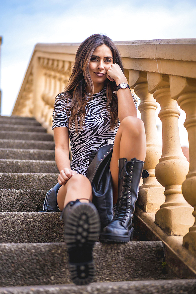 Lifestyle, look of a pretty brunette sitting on stairs with black boots and a black and white dress