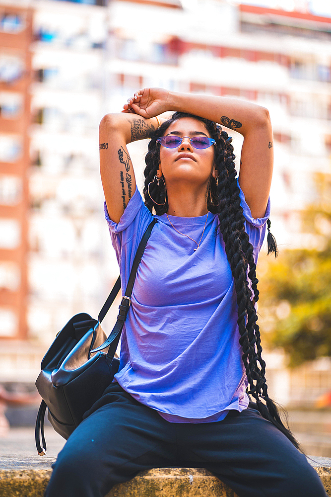 Lifestyle, young dark-skinned woman with long braids and purple glasses sitting on a bench in the park, with buildings in the background