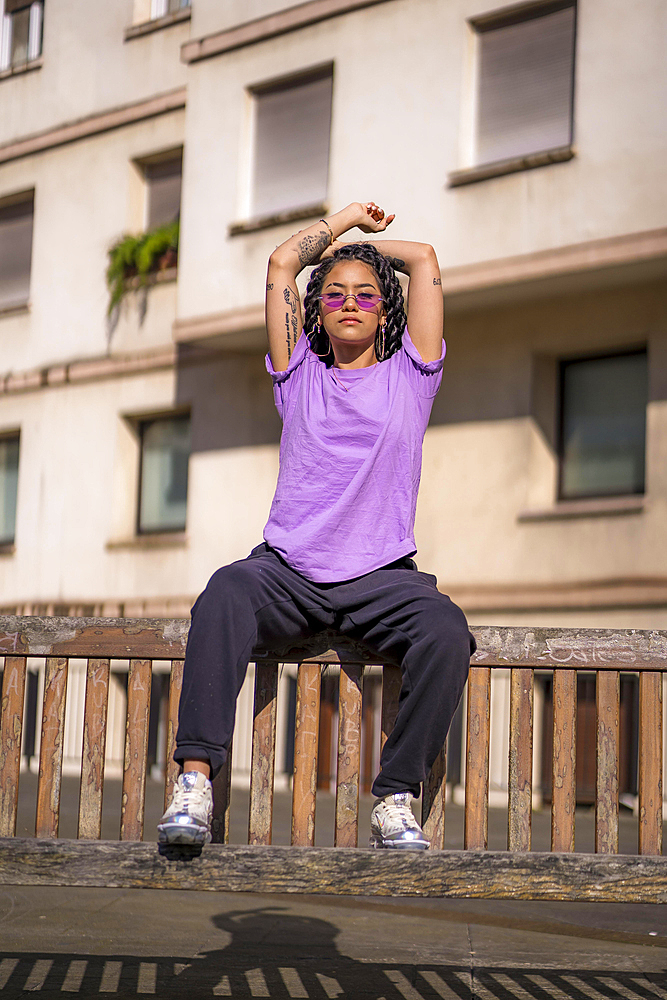 Young dark-skinned woman with long braids in purple glasses sitting on a bench in the park, with buildings in the background. With arms up