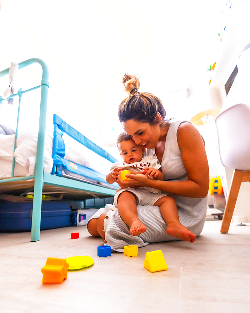 Young Caucasian mother playing with her in the room with toys. Baby less than a year learning the first lessons of her mother. Mother playing with her son and hugging him lovingly