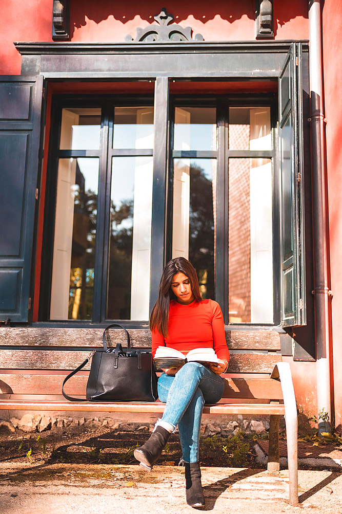 Lifestyle. Young Caucasian brunette in a red shirt sitting on a bench in the park reading a book. Vertical photo