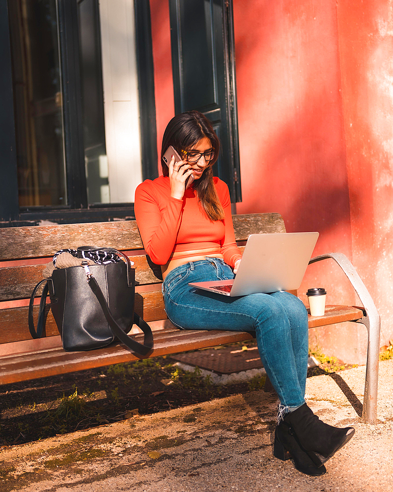 Lifestyle. Young caucasian brunette in a red shirt sitting on a bench in the park with a take away coffee. Talking on the phone and working with the computer