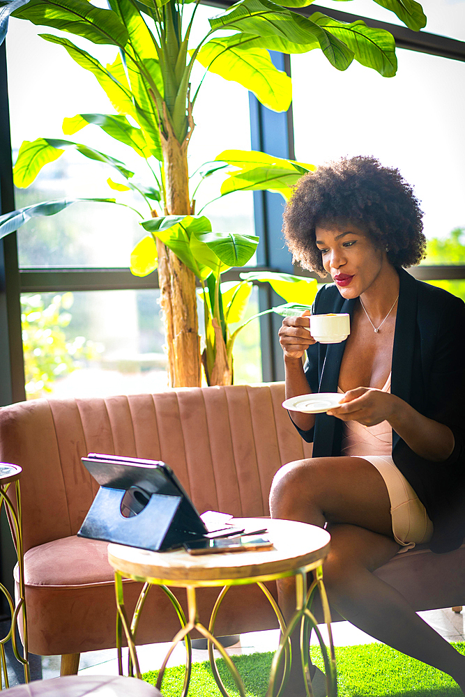 Black skinned girl with afro hair wearing a pink dress and black jacket, having a coffee and making a video call from work very happy. Sitting on a pink sofa, young enterprising woman in a hotel