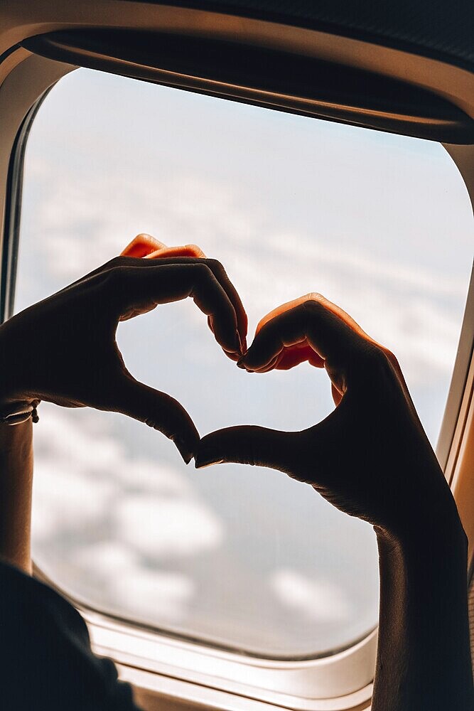 Female hand in heart shape Airplane flight. View from the window of the plane. Airplane, Aircraft. Vacation love travel concept Traveling by air. Airplane Window View Above the Clouds Amazing golden fluffy clouds moving softly on the sky aerial view