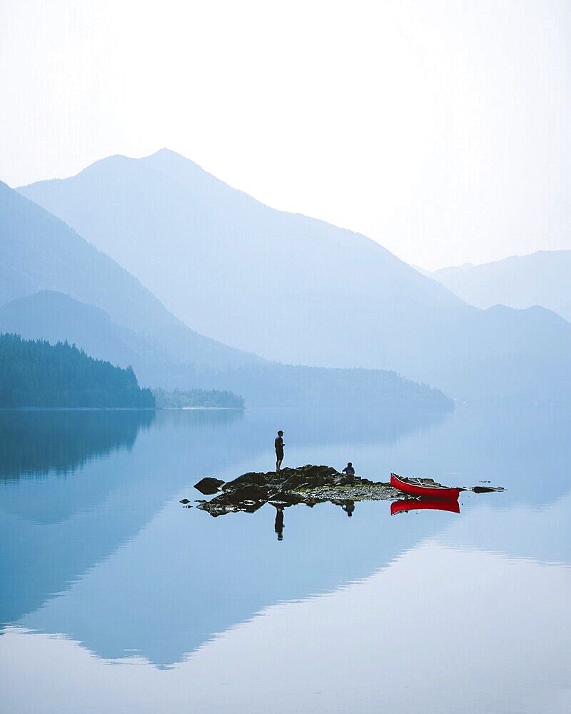 Person standing on a small island in a misty lake surrounded by mountains, with a red canoe nearby, Slocan Lake, British Columbia, Canada, North America