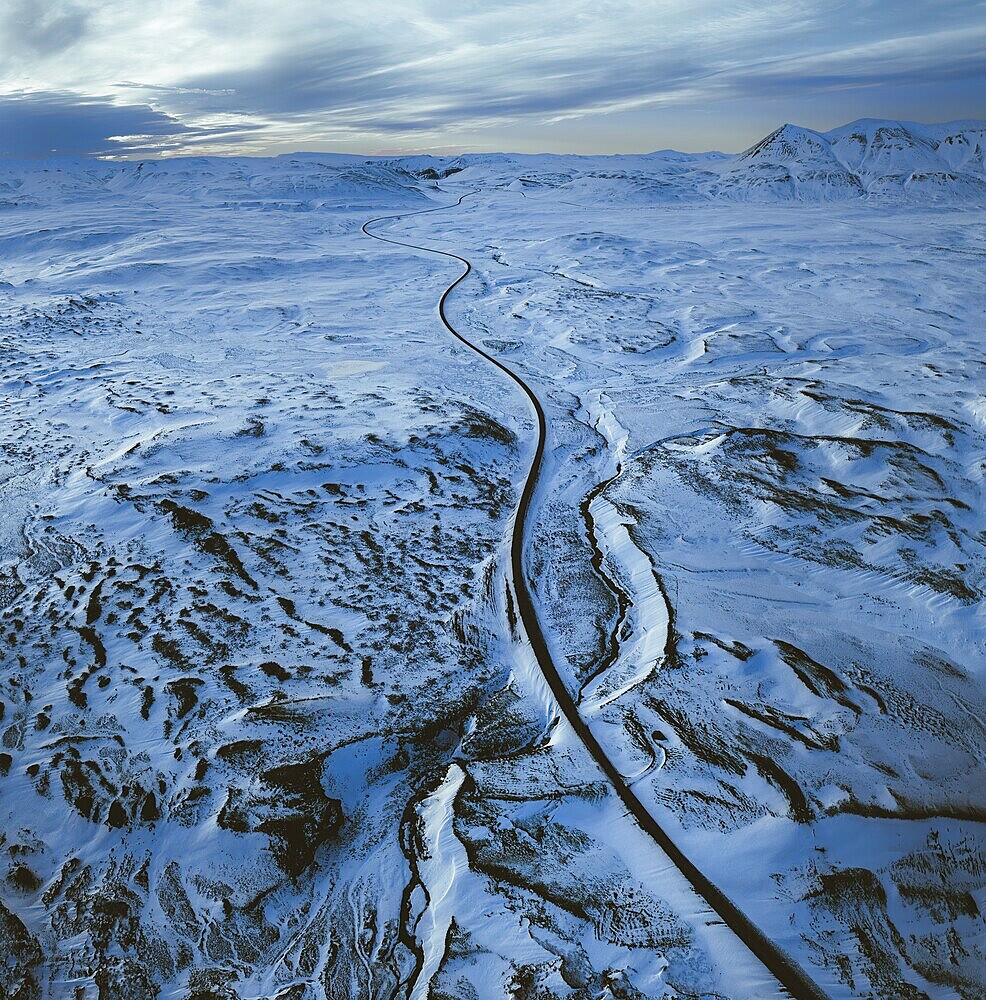 Aerial view of a long road winding through a vast snowy landscape with distant mountains under a cloudy sky, North, Iceland, Europe