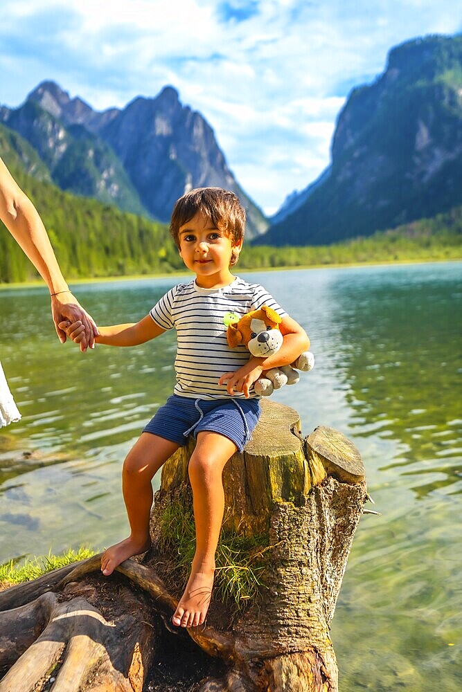 Young boy holding a plush toy and his mother's hand while sitting on a tree stump by lake dobbiaco, surrounded by the dolomites